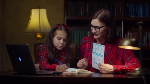 Chica de la escuela haciendo deberes de matemáticas con la joven madre de 30 años en casa. Un niño mirando ecuaciones matemáticas y anotando en un cuaderno. Proceso de educación en el hogar. Aspecto familiar mamá e hija . — Vídeos de Stock