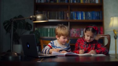 Evde online ödev yapan iki anaokulu çocuğu. Kardeşler dizüstü bilgisayara bakarak kalemlerle birlikte yazıyorlar. Çevrimiçi ev eğitimi süreci. 