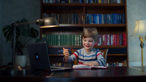 Προσχολικός τύπος κάνει online σχολική εργασία με φορητό υπολογιστή. Παιδί κάθεται στο ξύλινο γραφείο στο σπίτι και σπουδάζει online, γράφοντας με στυλό στο βιβλίο ασκήσεων. — Αρχείο Βίντεο