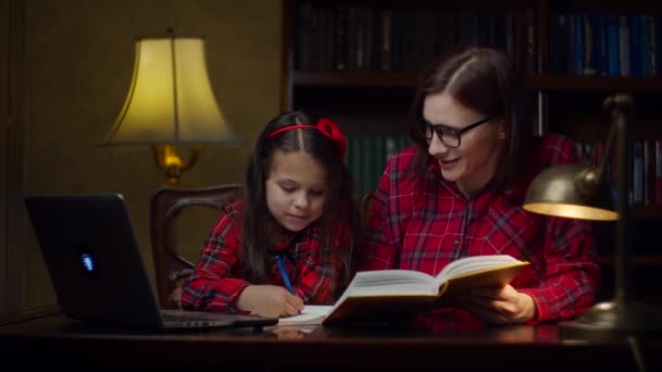 Schoolmeisje maakt huiswerk met jonge dertiger moeder thuis. Een jongen die naar het leerboek kijkt en opschrijft in een notitieboekje. Thuisonderwijs. Familie kijken mam en dochter. — Stockvideo