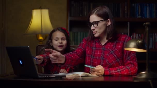 30 'lu yaşlardaki genç annesiyle ev ödevi yapan liseli kız evde cep telefonu kullanıyor. Çocuk dizüstü bilgisayara bakıyor ve not defterine yazıyor. Çevrimiçi eğitim süreci. Aile bakımı anne ve kız. — Stok video