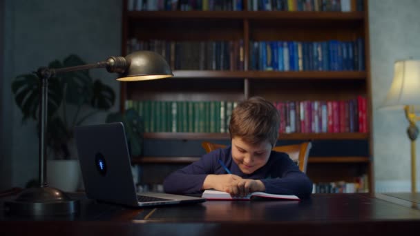 Uczeń odrabiający lekcje online z laptopem w domu. Dzieciak siedzi przy drewnianym biurku i uczy się online, zapisuje długopisem w zeszycie ćwiczeń. — Wideo stockowe