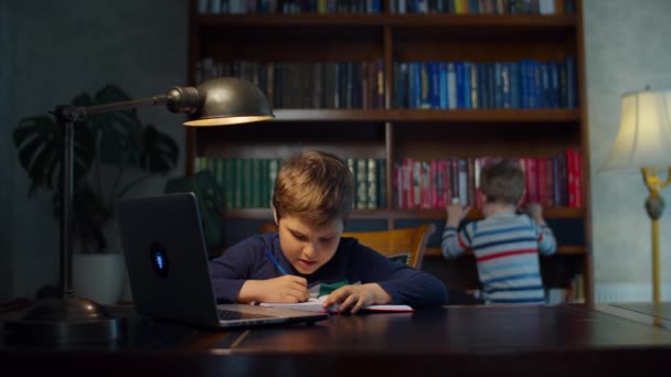 Niño de la escuela haciendo tarea de la escuela en línea con el ordenador portátil en casa. El hermano menor se queda con el libro. Niño sentado en el escritorio de madera y estudiando en línea, escribiendo con pluma en el libro de ejercicios . — Vídeo de stock