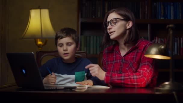 School jongen maakt online huiswerk met jonge dertiger moeder thuis. Een jongen die naar een laptop kijkt en opschrijft in een notitieboekje. Online onderwijsproces thuis. — Stockvideo