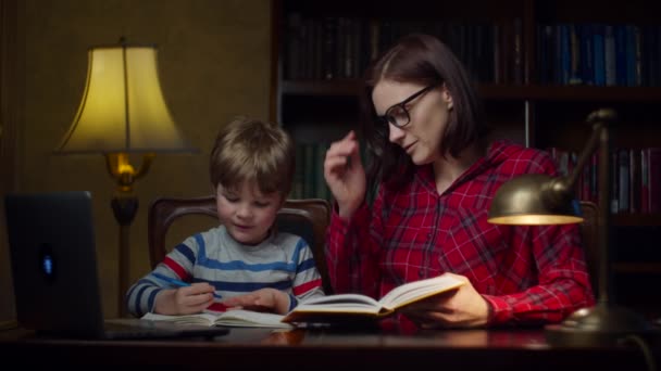 Niño preescolar haciendo tarea en línea con la madre joven de 30 años en casa. Un chico mirando un libro de texto y anotando en un cuaderno. Home proceso de educación en línea . — Vídeo de stock