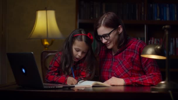 Schoolmeisje maakt huiswerk met jonge dertiger moeder thuis. Een jongen die naar een laptop kijkt en opschrijft in een notitieboekje. Online onderwijsproces. Familie kijken mam en dochter. — Stockvideo