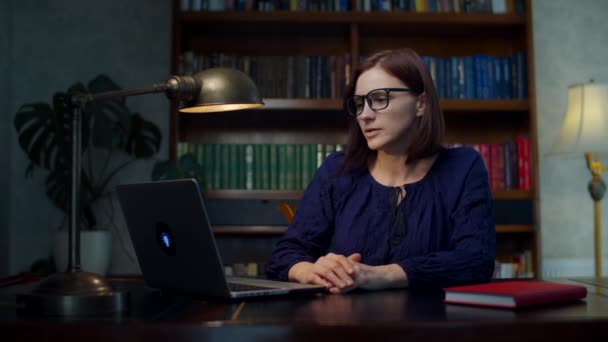 Brunette femme des années 30 dans des lunettes parlant à un ordinateur portable assis à la table en bois à la maison avec bibliothèque. Travailler de la femme à la maison en utilisant un appel vidéo par ordinateur portable . — Video