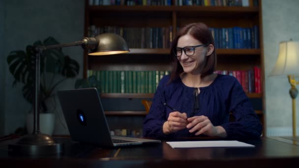 Feliz morena mujer de 30 años en gafas trabajando desde casa firmando documentos de papel sentado en la mesa de madera con estantería. Mujer sonriente trabaja desde casa . — Vídeo de stock