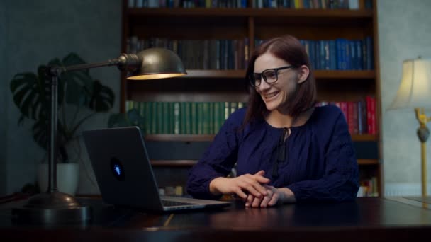 Brunett 30-tals kvinna i glasögon pratar med bärbar dator sitter vid träbord hemma med bokhylla. Arbeta hemifrån kvinna med hjälp av videosamtal med bärbar dator. — Stockvideo
