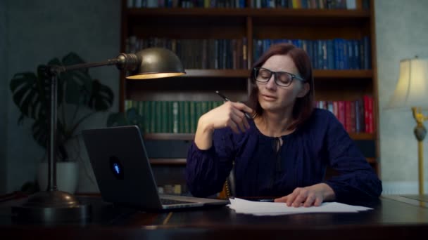 Расстроенная женщина 30-х годов в очках, работающая из дома, подписывая бумажные документы, сидя за деревянным столом. Несчастная женщина смотрит на ноутбук . — стоковое видео