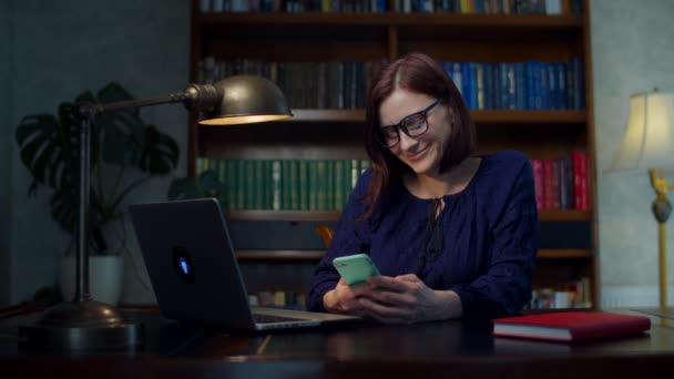 Jovens 30 anos trabalhando de mulher em casa em óculos usando telefone celular sentado à mesa de madeira com laptop em casa. Mulher sorridente com célula . — Vídeo de Stock