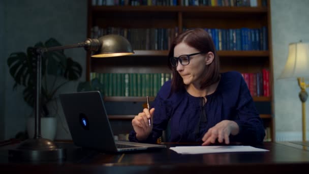 Brünette Frau in den 30er Jahren mit Brille, die von zu Hause aus Papierdokumente signiert, sitzt am Holztisch mit Bücherregal. Weibchen mit Laptop checkt Dokumente zu Hause. — Stockvideo