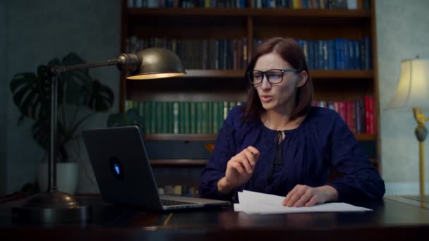 Verontrustende vrouw uit de 30 in een bril die thuis papieren documenten signeert zittend aan een houten tafel. Ongelukkig vrouwtje op zoek naar laptop. — Stockvideo