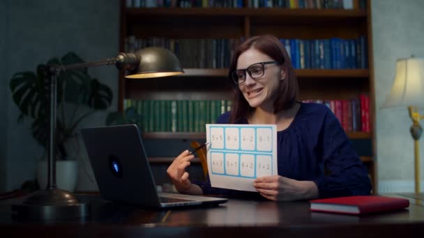 Eine junge Lehrerin in den Dreißigern mit Brille spricht und zeigt ein Blatt Papier mit einfachen mathematischen Gleichungen, das zu Hause auf den Laptop schaut. Online-Bildungsprozess. Arbeit von zu Hause aus. — Stockvideo