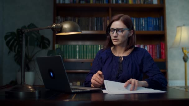 Brünette Frau in den 30er Jahren mit Brille, die von zu Hause aus Papierdokumente signiert, sitzt am Holztisch mit Bücherregal. Weibchen mit Laptop checkt Dokumente zu Hause. — Stockvideo