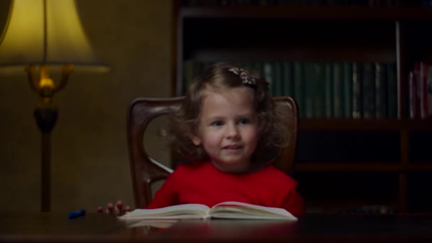 Schattig kleuter meisje in rode jurk maken huiswerk schrijven in notitieboekje met pen zitten aan houten tafel 's avonds thuis. Thuisonderwijs. — Stockvideo