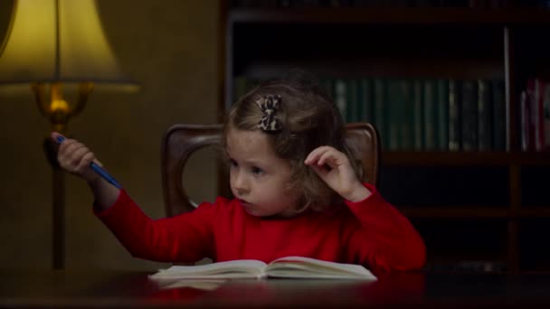 Мила дівчина дошкільного віку в червоній сукні робить домашнє завдання, пишучи в блокноті з ручкою, сидячи за дерев'яним столом ввечері вдома. Процес домашньої освіти . — стокове відео