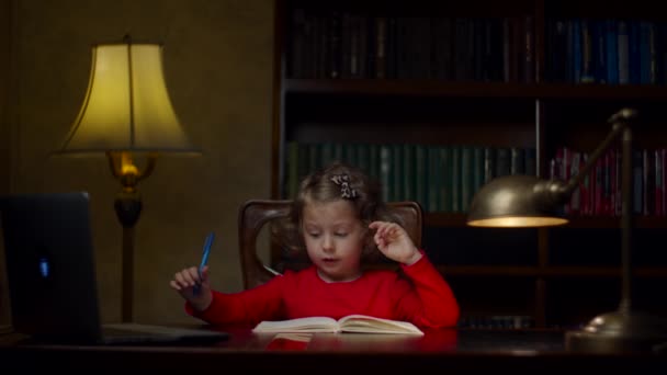 Cute preschool girl in red dress making homework writing in notebook with pen sitting at wooden table at the evening at home. Home education process. — Stock Video