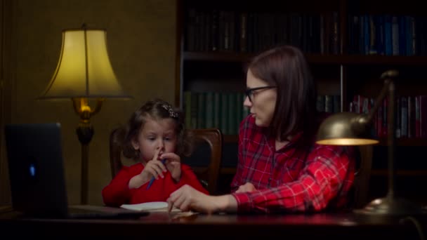 Kleutermeisje maakt online huiswerk met jonge dertiger moeder thuis. Schattig klein meisje dat opschrijft in het notitieboekje. Thuis online onderwijs proces. Moeder en dochter in rode jurken. — Stockvideo