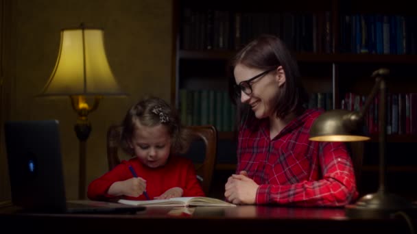 Förskola flicka gör online läxor med unga 30-talet mor hemma. Söt liten flicka som skriver ner i anteckningsboken. Hemundervisning på nätet. Mamma och dotter i röda klänningar. — Stockvideo