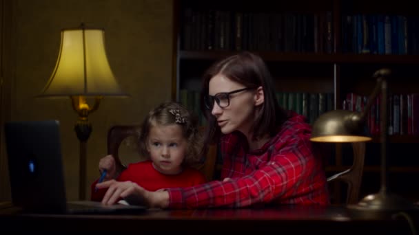 Kleutermeisje maakt online huiswerk met jonge moeder thuis. Leuk klein meisje en een vrouw van in de 30 die met haar handen zwaait naar een laptop. Online onderwijsproces. Moeder en dochter in rode jurken. — Stockvideo