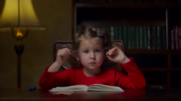 Retrato de linda niña preescolar en vestido rojo sonriendo mirando a la cámara sentada en la mesa de madera por la noche en casa en el interior clásico . — Vídeo de stock