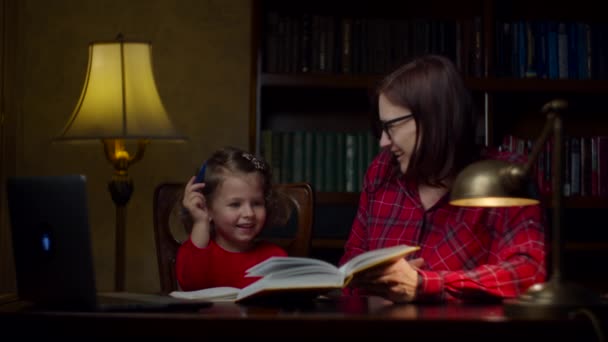Дошкільна дівчинка робить домашнє завдання з матір'ю 30-х років. Милий хлопчина дивиться на підручник і записує в зошит. Процес домашньої освіти. Мама з дочкою в червоних сукнях.. — стокове відео