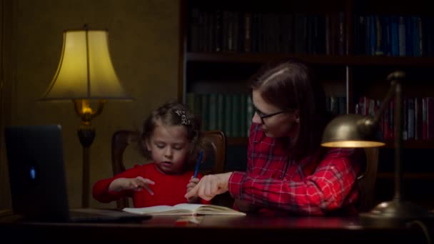 Een kleuter die huiswerk maakt met een moeder van in de dertig. Schattig klein meisje dat opschrijft in het notitieboekje. Thuisonderwijs. Moeder en dochter in rode jurken. — Stockvideo