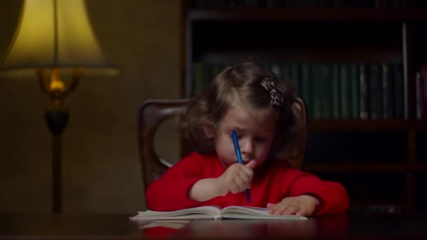 Linda niña preescolar en vestido rojo haciendo tarea de escritura en cuaderno con pluma sentado en la mesa de madera en la noche en casa. Proceso de educación en el hogar . — Vídeos de Stock