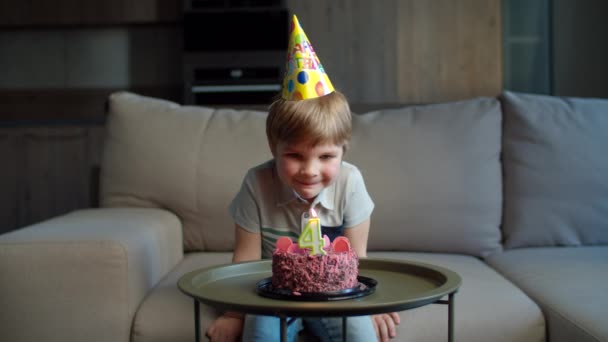 Niño preescolar en sombrero de cumpleaños soplando vela con el número 4 en pastel de cumpleaños de chocolate solo en casa. Niño celebra 4 años de cumpleaños en casa solo . — Vídeos de Stock