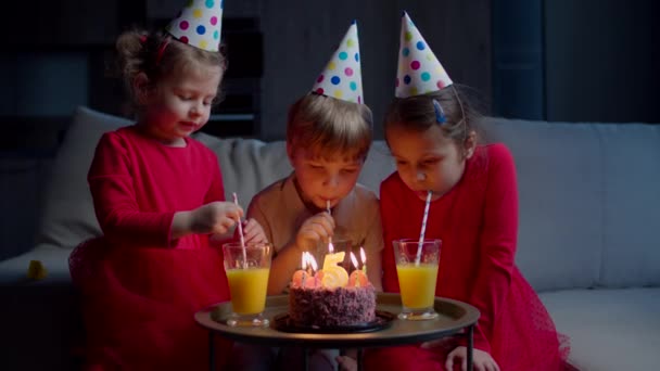 Trois enfants dans des chapeaux d'anniversaire buvant du jus de fruits avec de la paille à la maison. Frères et sœurs célèbrent leur anniversaire avec un gâteau au chocolat aux bougies assis sur le canapé dans le salon . — Video
