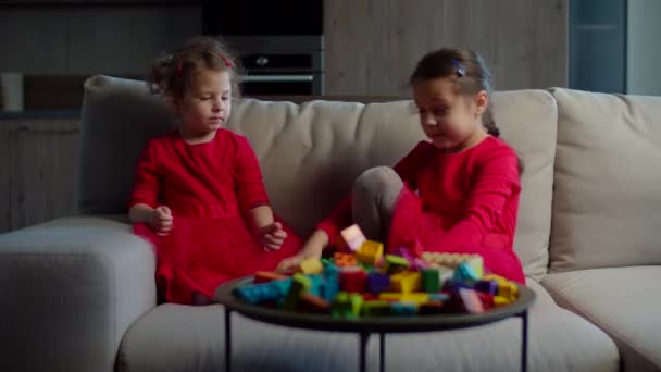 Δύο παιδιά που παίζουν με μπλοκ έγχρωμων κατασκευαστών κάθονται στον καναπέ στο σπίτι. Αδερφές με τα ίδια ρούχα παίζουν μαζί σε αργή κίνηση. Δύο κορίτσια με κόκκινα φορέματα.. — Αρχείο Βίντεο