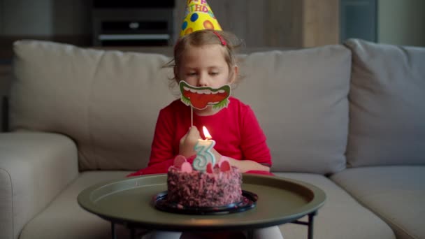 Børnehave pige i fødselsdagshat blæser ud stearinlys med nummer 3 på chokolade fødselsdagskage alene hjemme. Kid fejrer fødselsdag hjemme alene . – Stock-video