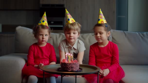 Drie kinderen met verjaardagshoed vieren hun verjaardag met chocoladetaart met kaarsen thuis. Broer blaast verjaardagskaarsen uit op taart met zussen zittend op de bank in de woonkamer. — Stockvideo