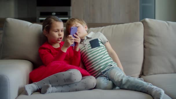 School girl using mobile phone application with brother watching sitting on couch at home. Siblings using gadget together. — Stock Video