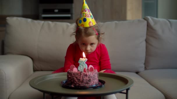 Niña preescolar en sombrero de cumpleaños soplando vela con el número 3 en pastel de cumpleaños de chocolate solo en casa. El niño celebra su cumpleaños solo en casa. . — Vídeo de stock