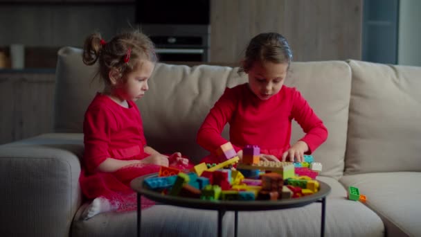 İki çocuk evdeki kanepede oturan renkli bloklarla oynuyorlar. Aynı kıyafetleri giyen kardeşler ağır çekimde birlikte oynuyorlar. Kırmızı elbiseli iki kız.. — Stok video