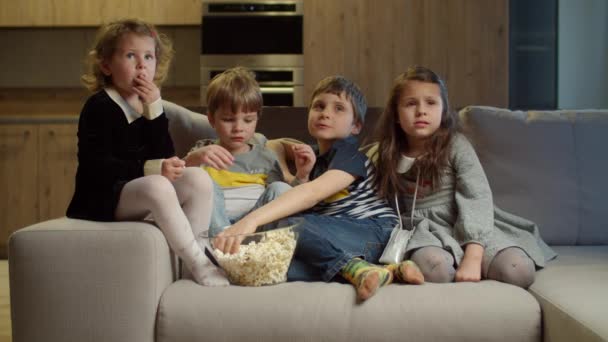 Grupo de quatro crianças assistindo TV, comendo pipocas sentadas no sofá em casa. Duas meninas e dois meninos assistindo filme emocionante em casa. Irmãos no sofá . — Vídeo de Stock