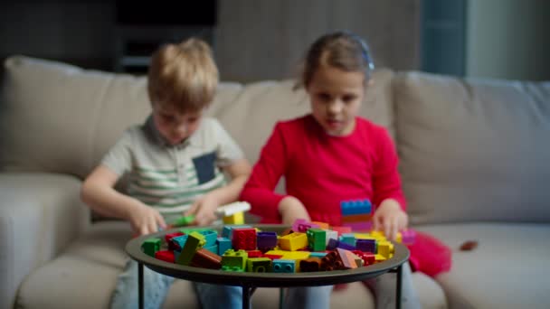 Duas crianças brincando com blocos de construtor de cores sentadas no sofá em casa. Menina em vestido vermelho e construção menino com construtor. Os irmãos brincam juntos. Foco no construtor . — Vídeo de Stock