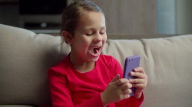 Kırmızı elbiseli kız cep telefonu uygulaması kullanıyor ve evdeki koltukta oturan cep kamerasına komik suratlar yapıyor. Küçük kız kardeş onu mutlu etmeye geliyor.. 