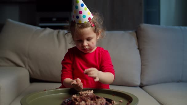 Linda niña en sombrero de fiesta comiendo pastel de chocolate de cumpleaños con las manos en casa. Cumpleaños niño disfrutando de comer dulces solo en casa . — Vídeos de Stock
