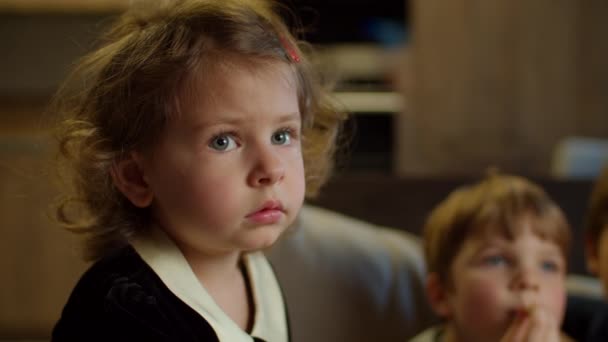 Linda chica viendo la televisión, comiendo palomitas de maíz sentado en el sofá en casa. Dos niños viendo películas emocionantes en casa. Hermanos en el sofá. De cerca. — Vídeos de Stock