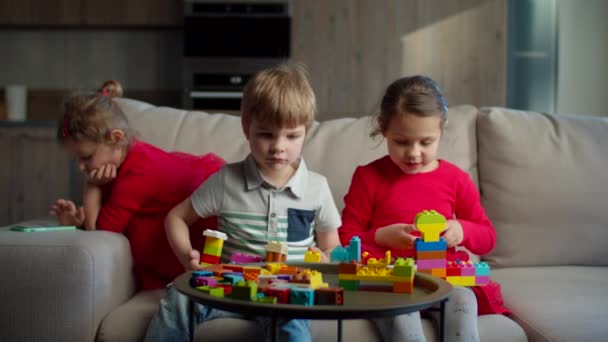 Dos niños jugando con bloques de construcción de color y una niña pequeña usando aplicaciones de teléfono móvil sentados en el sofá en casa. Hermanos jugando juntos . — Vídeos de Stock