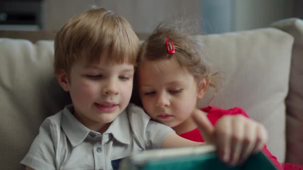 Przedszkolak i dziewczynka za pomocą tabletu siedzi na kanapie w domu. Dzieci korzystające z aplikacji mobilnej na tablecie razem. — Wideo stockowe