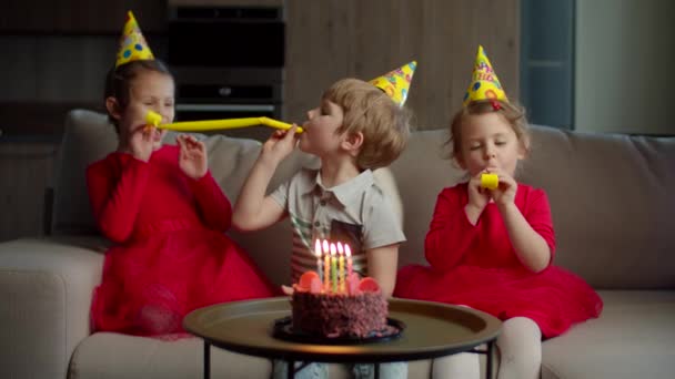 Drei glückliche Kinder mit Geburtstagsmützen blasen sich zu Hause Geburtstagspfeifen zu. Geschwister feiern Geburtstag mit Schokokuchen bei Kerze auf Couch im Wohnzimmer. — Stockvideo