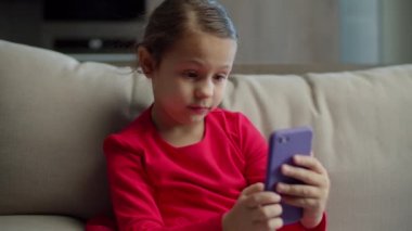 Kırmızı elbiseli kız cep telefonu uygulaması kullanıyor ve evdeki koltukta oturan cep kamerasına komik suratlar yapıyor.. 