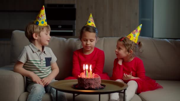 Drei Kinder mit Geburtstagsmützen feiern Geburtstag mit Schokokuchen und Kerzen zu Hause. Mädchen bläst Geburtstagskerzen auf Kuchen aus, Geschwister sitzen auf Couch im Wohnzimmer. — Stockvideo