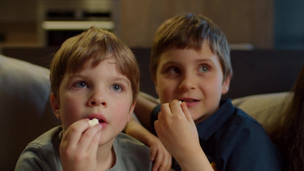 Dois irmãos assistindo TV, comendo pipocas sentados no sofá em casa. Dois meninos assistindo filme emocionante em casa. Irmãos no sofá . — Vídeo de Stock