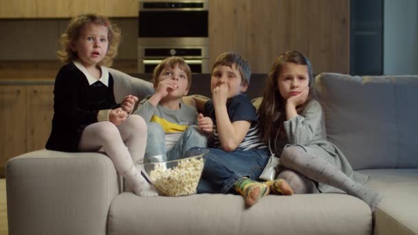 Grupa czworga dzieci ogląda telewizję, je popcorn siedząc na kanapie w domu. Dwie dziewczyny i dwóch chłopców oglądających ekscytujący film w domu. Rodzeństwo na kanapie. — Wideo stockowe
