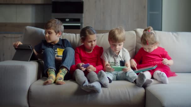 Ομάδα τεσσάρων παιδιών μικτής ηλικίας που χρησιμοποιούν gadgets κάθονται μαζί στον καναπέ στο σπίτι. Αδέρφια που παίζουν και παρακολουθούν σε απευθείας σύνδεση σε κινητά τηλέφωνα και υπολογιστές tablet. Δύο αγόρια και δύο κορίτσια με συσκευές. — Αρχείο Βίντεο